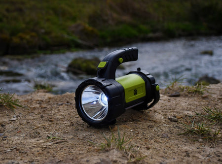A Six Peaks Multi-function Searchlight Lantern by Six Peaks, in green and black, is switched on and resting on sandy ground near a river, with grass and water visible in the background.
