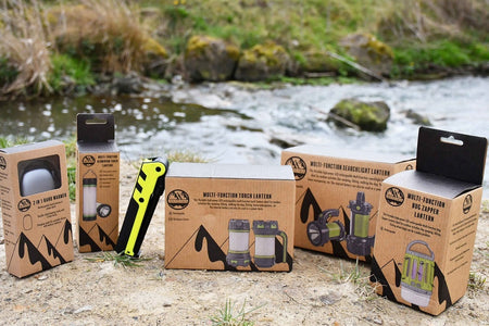 Six Peaks Multi-function Searchlight Lanterns by Six Peaks, packed in boxes, are shown on sandy ground near a stream with grass and rocks in the background.
