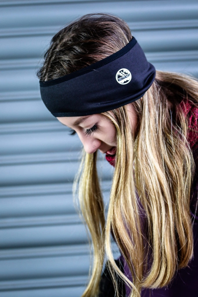 A person with long blonde hair wears the Six Peaks Winter Ear Warmer, a black, fleece-lined, wind-resistant headband by Six Peaks with a small mountain patch, standing before a grey metal shutter.