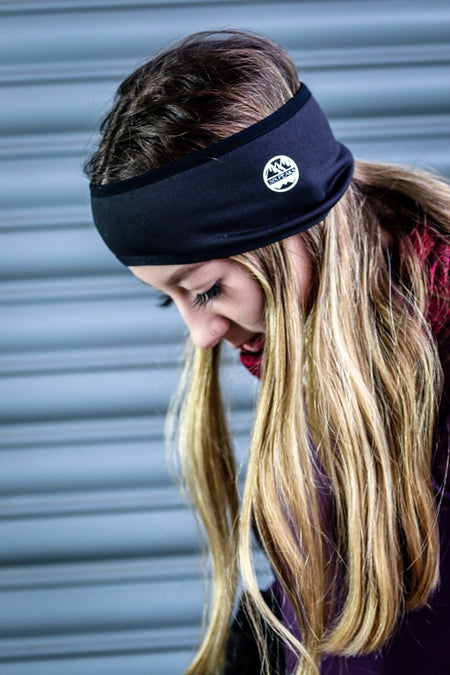 A person with long blonde hair wears the Six Peaks Winter Ear Warmer, a black, fleece-lined, wind-resistant headband by Six Peaks with a small mountain patch, standing before a grey metal shutter.