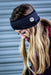 A person with long blonde hair wears the Six Peaks Winter Ear Warmer, a black, fleece-lined, wind-resistant headband by Six Peaks with a small mountain patch, standing before a grey metal shutter.