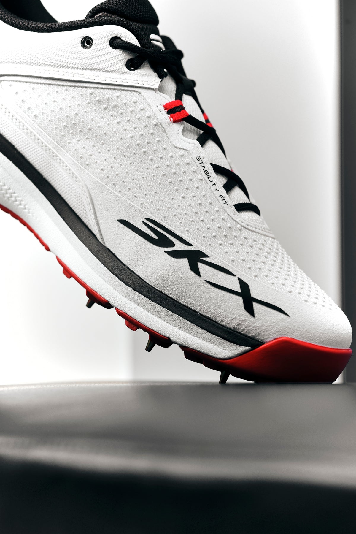 A close-up of the Skechers Cricket Elite Cricket Shoe shows black and red accents, black laces, SKX branding, an Arch Fit insole, and sole spikes. The white shoe by Skechers is angled upward against a bright background.