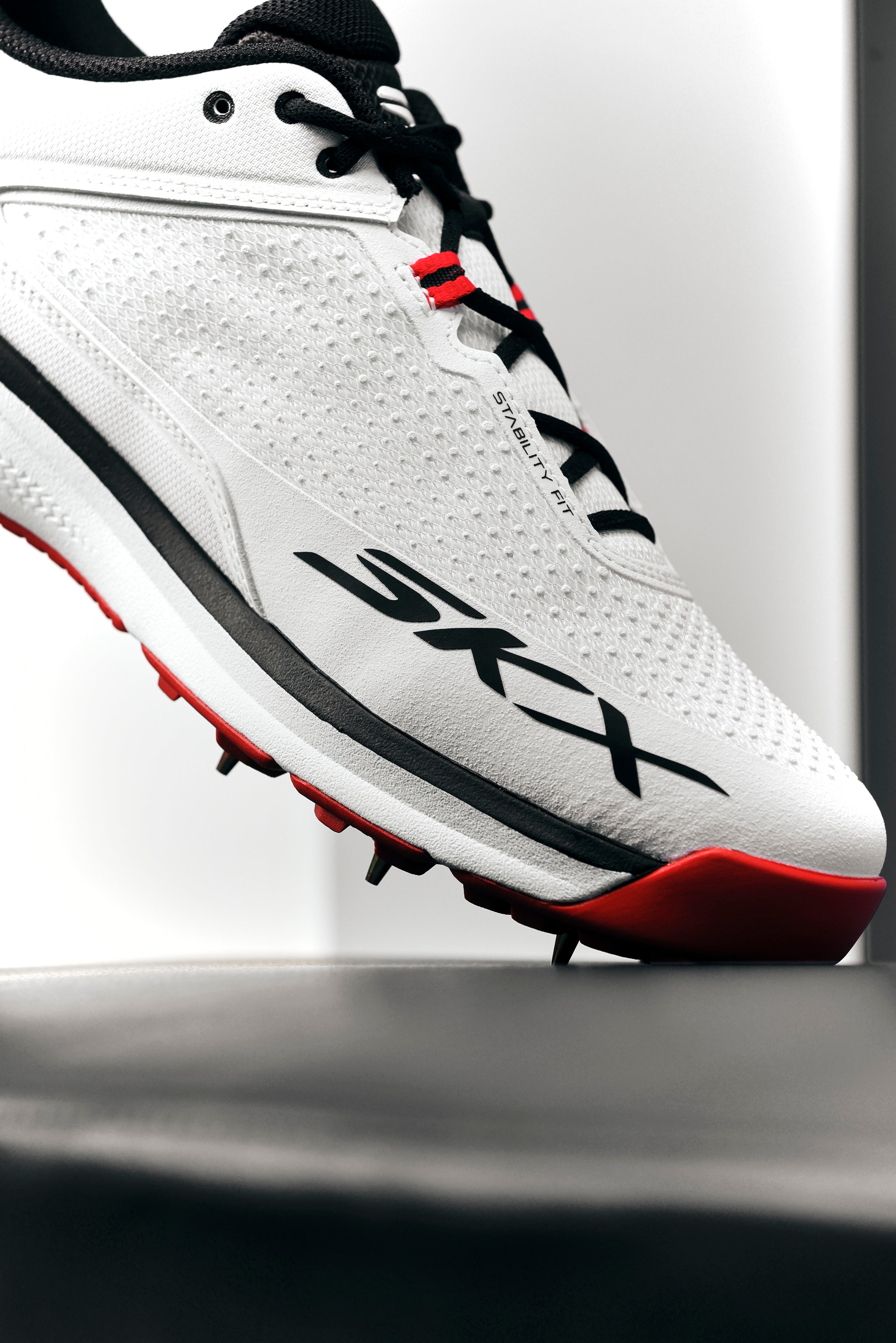 A close-up of the Skechers Cricket Elite Cricket Shoe shows black and red accents, black laces, SKX branding, an Arch Fit insole, and sole spikes. The white shoe by Skechers is angled upward against a bright background.