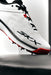 A close-up of the Skechers Cricket Elite Cricket Shoe shows black and red accents, black laces, SKX branding, an Arch Fit insole, and sole spikes. The white shoe by Skechers is angled upward against a bright background.
