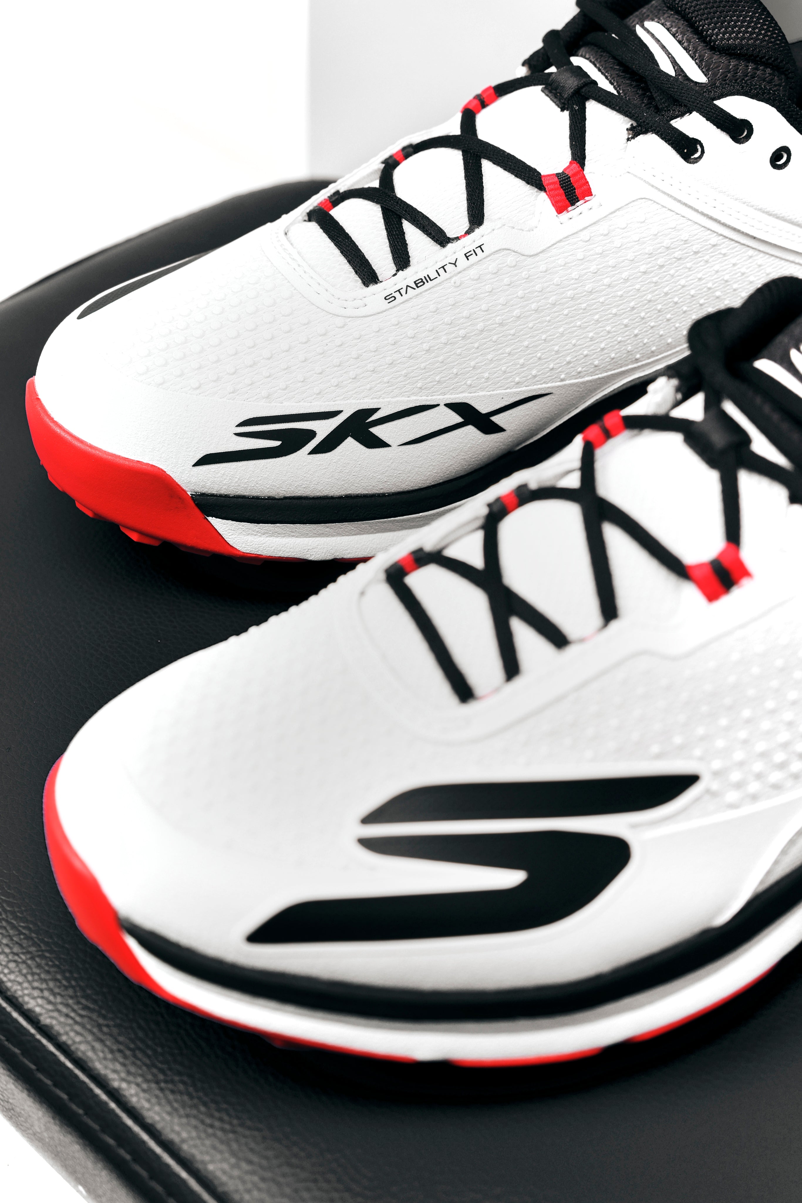 A pair of white Skechers Cricket Elite Cricket Shoes with black laces, red accents, and an Arch Fit insole rest on a black surface.