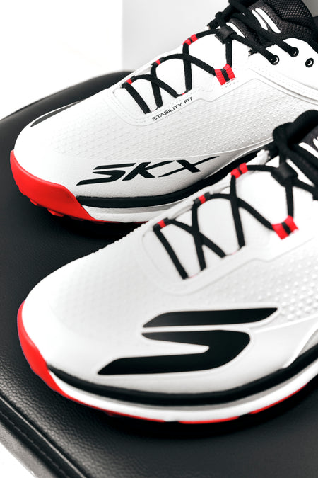 A pair of white Skechers Cricket Elite Cricket Shoes with black laces, red accents, and an Arch Fit insole rest on a black surface.