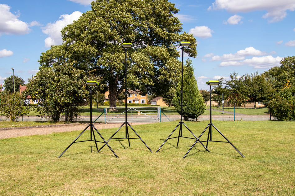 Four Ritelite Sports Lite Portable Floodlights stand on a grassy field with trees, a fence, and houses in the background under a blue sky with scattered clouds.