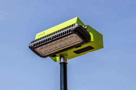 A Ritelite Sports Lite Portable Floodlight, with a bright green rectangular design and multiple visible LED bulbs, is mounted on a black pole against a clear blue sky.
