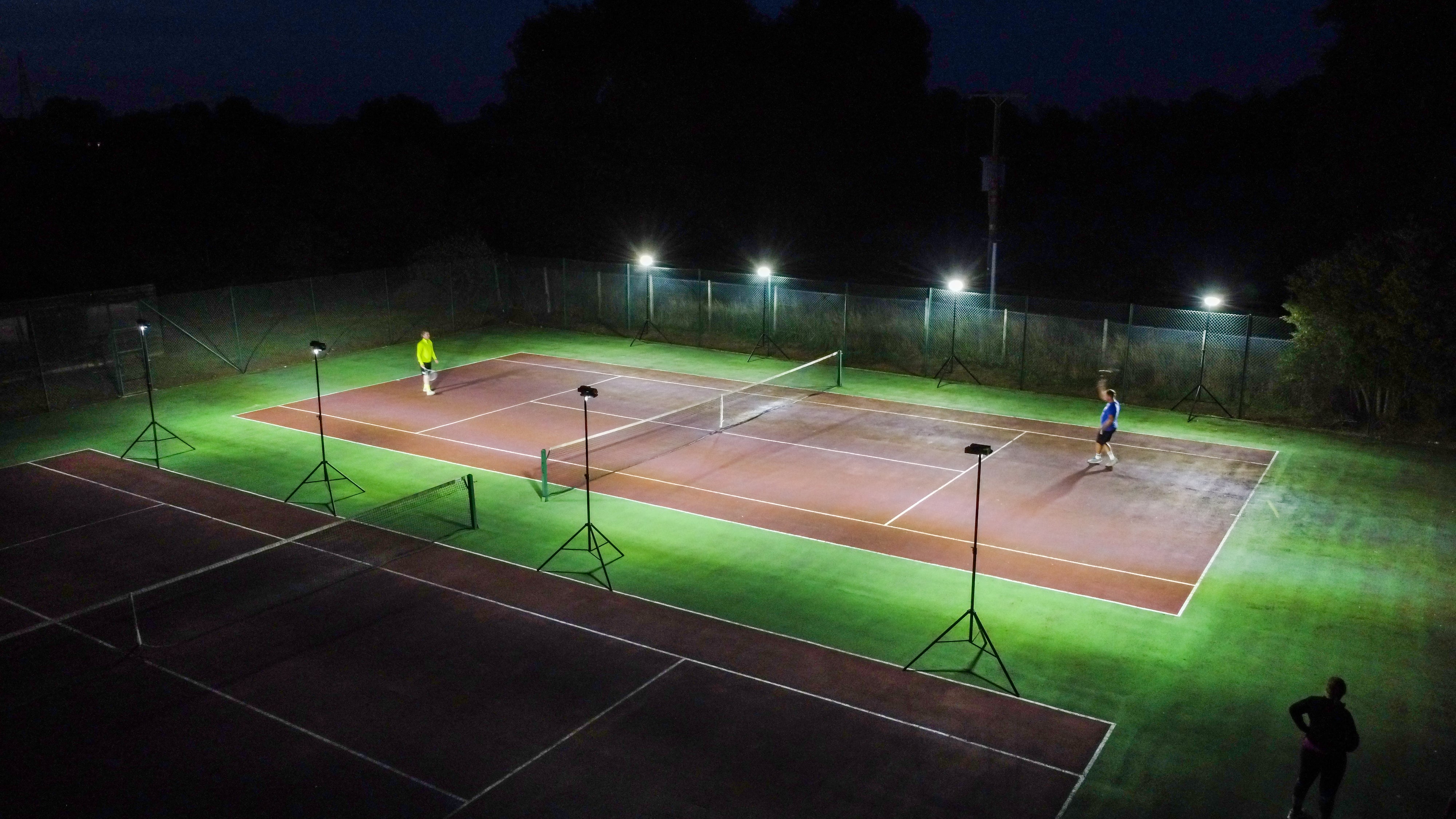 Aerial view of two people playing tennis at night on an outdoor court, illuminated by bright Ritelite Sports Lite Portable Floodlights. The surrounding area is dark and the court lines are clearly visible.