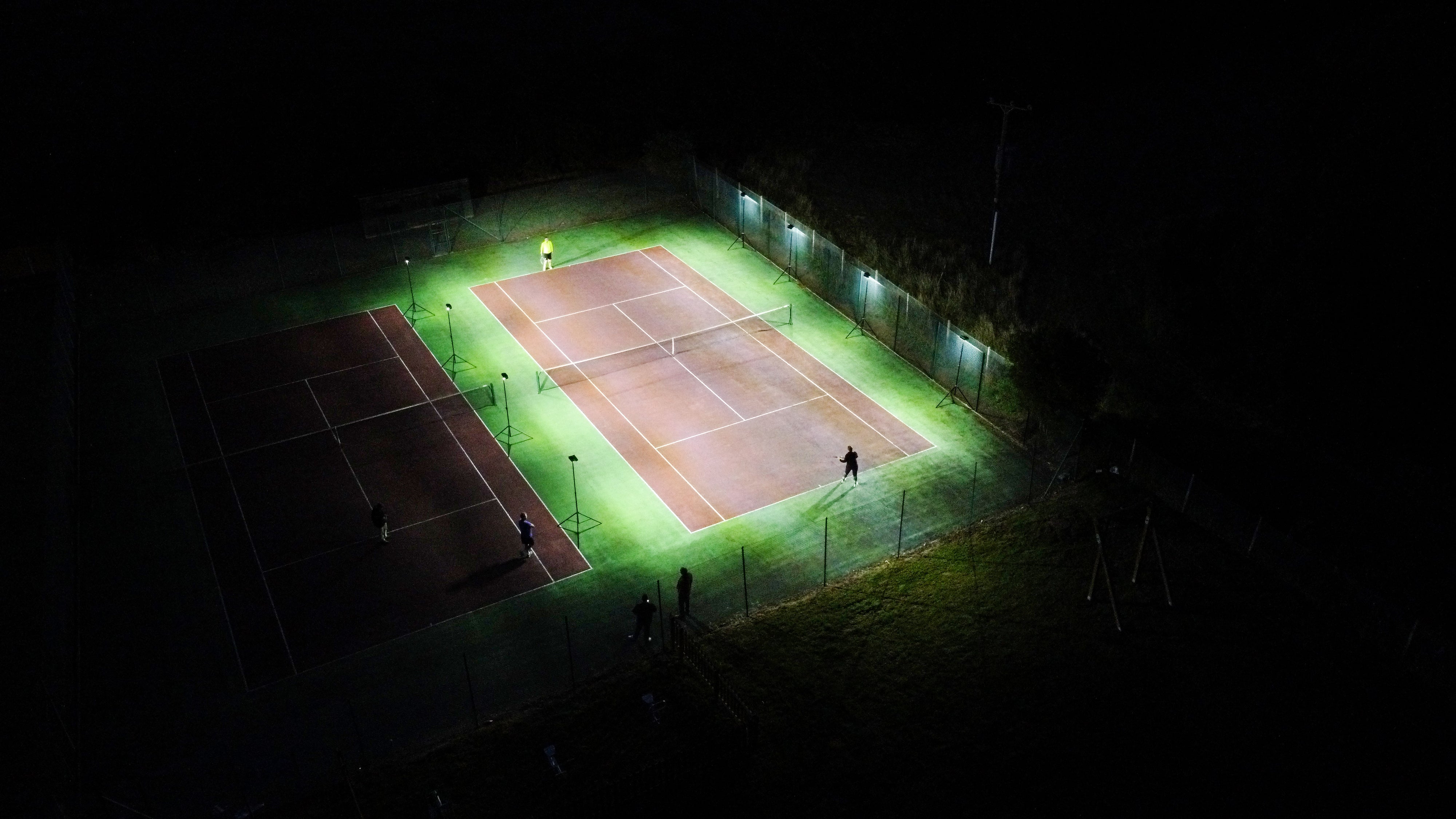 Aerial view of two adjacent tennis courts at night: one brightly lit by Ritelite Sports Lite Portable Floodlights with players on court, while the other remains mostly dark.