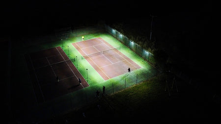 Aerial view of two adjacent tennis courts at night: one brightly lit by Ritelite Sports Lite Portable Floodlights with players on court, while the other remains mostly dark.