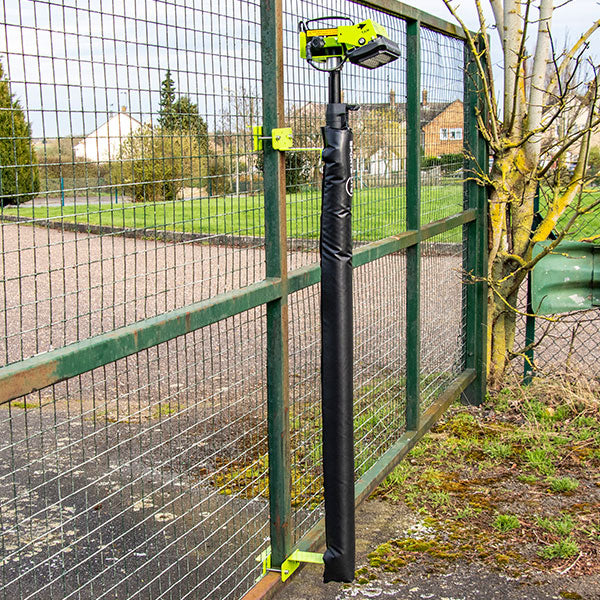 A green and black power tool hangs vertically on a wire mesh fence outdoors, secured with a black cover, a green bracket, and Ritelite Sports Lite Fence Kit Padding. Trees and houses are visible in the background.