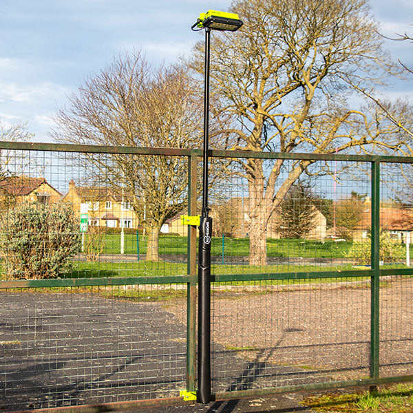 A tall black security camera is mounted on a pole with Ritelite Sports Lite Fence Kit Padding for player protection, attached to a metal gate in a park, with trees and houses in the background.