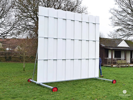 The Stuart Canvas Polypropylene Sightscreen, large and white with red wheels, stands on grass near a building and trees. A person is partially visible behind the cricket sightscreen.