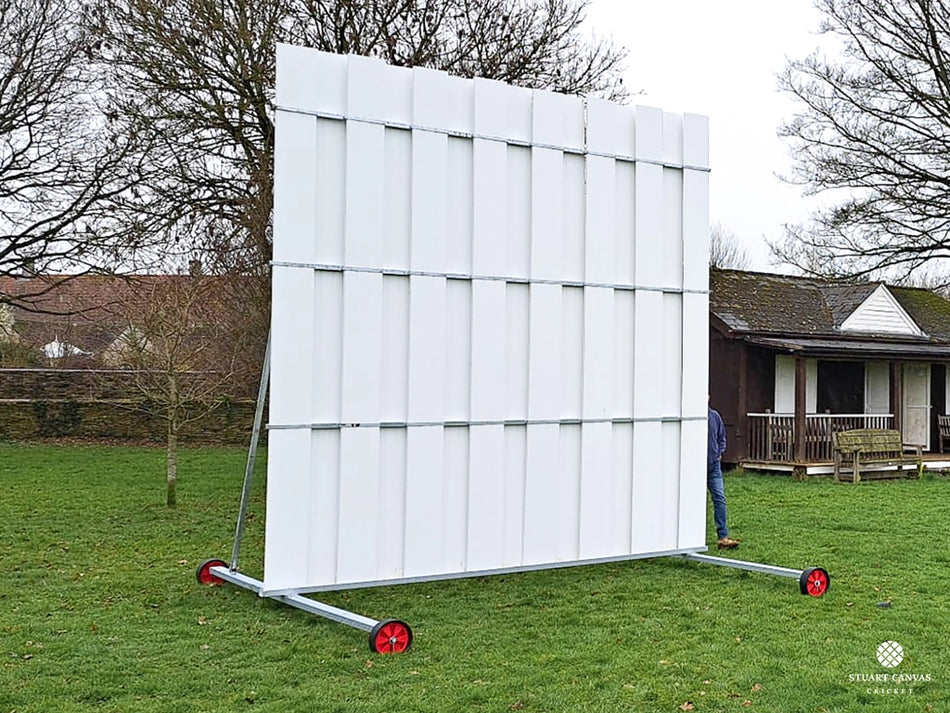 The Stuart Canvas Polypropylene Sightscreen, large and white with red wheels, stands on grass near a building and trees. A person is partially visible behind the cricket sightscreen.