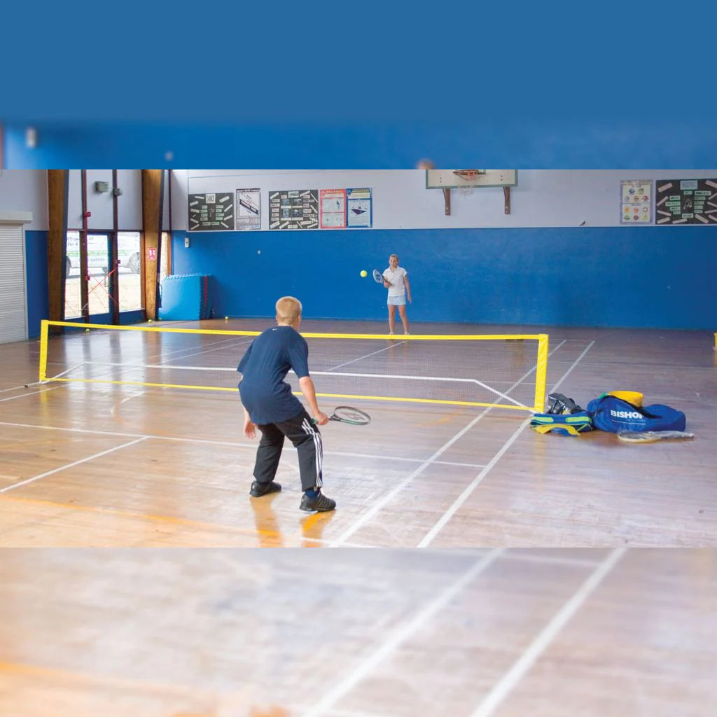 Two people play badminton on a wood-floored indoor court with blue walls, using the Bishop Sports Kwik Net by Bishop Sports. Sports bags and equipment are placed on the right side near the net.