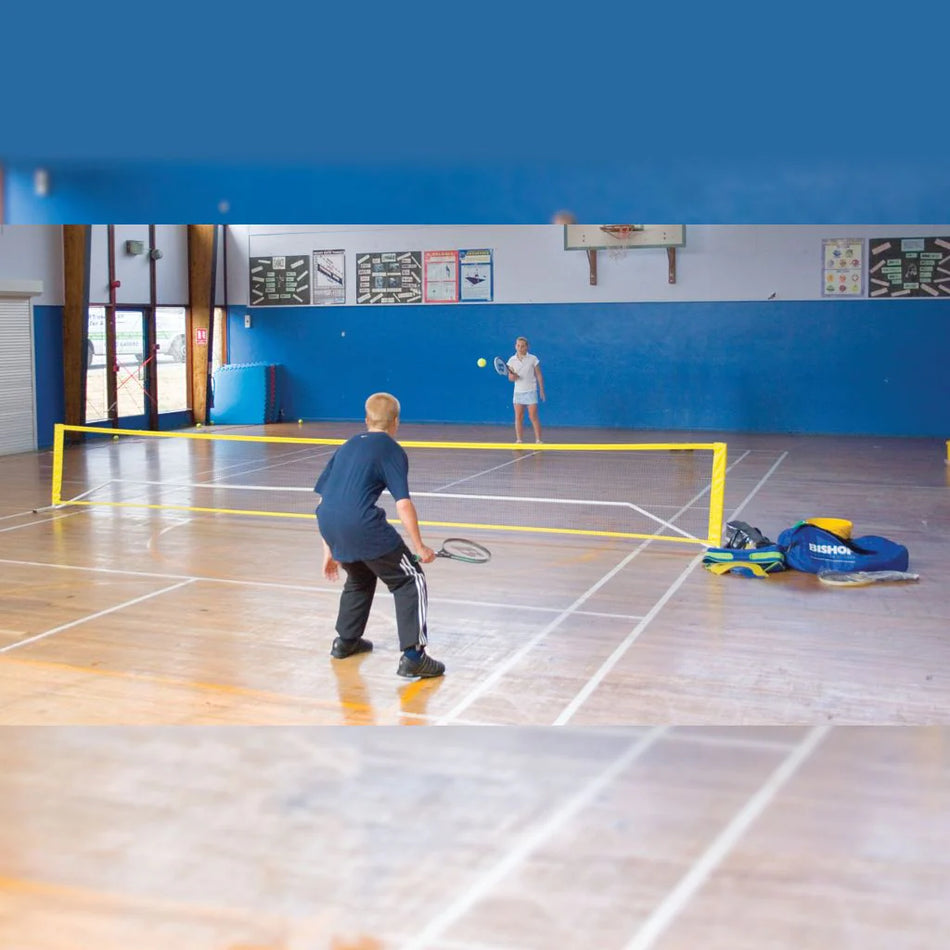 Two people play badminton on a wood-floored indoor court with blue walls, using the Bishop Sports Kwik Net by Bishop Sports. Sports bags and equipment are placed on the right side near the net.