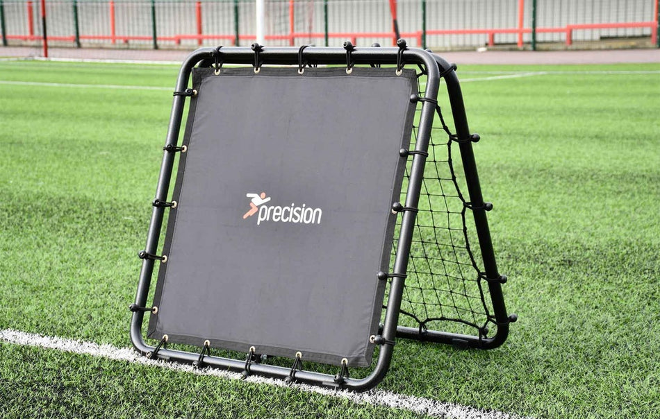The Reydon Sports Precision Pro Double Sided Rebounder, in black and printed with "precision," sits on green artificial turf near a white sideline, enclosed by a metal fence—ideal for skills training.