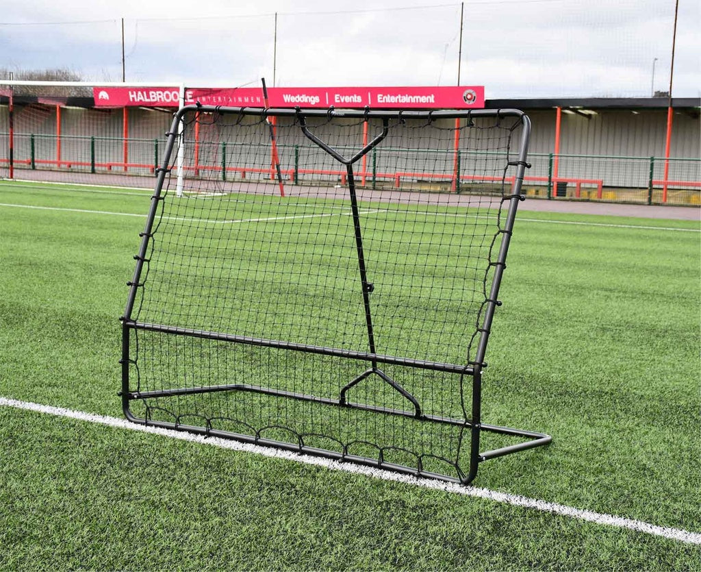 The Precision Training Precision Pro Two Angled Rebounder, a black metal net, is set on green artificial turf with empty stadium seats and a red and gray building in the background.