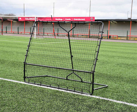 The Precision Training Precision Pro Two Angled Rebounder, a black metal net, is set on green artificial turf with empty stadium seats and a red and gray building in the background.