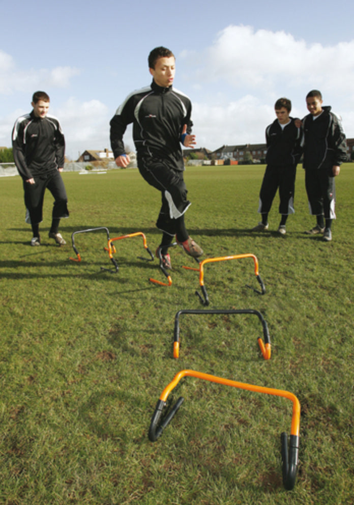 A young man in a black tracksuit runs and jumps over Precision Training Adjustable 6"/12" Hurdles Set on a grassy field, while three others in similar outfits watch in the background.