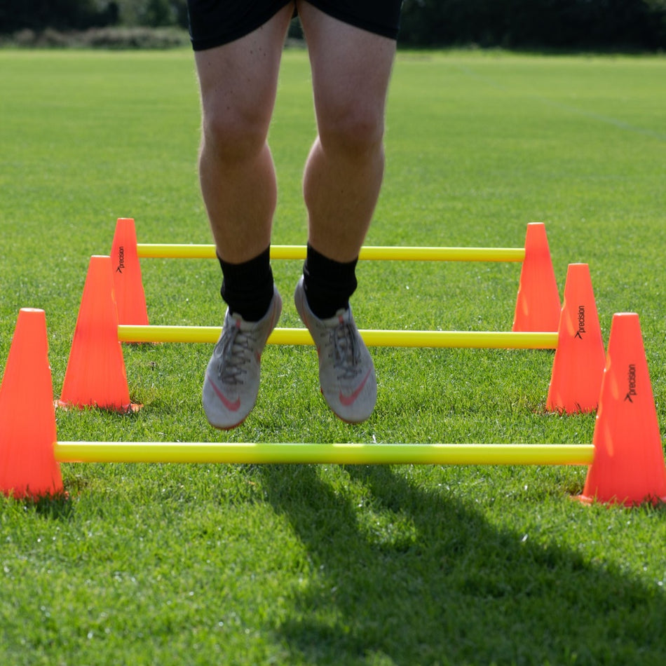 A person's lower legs and feet are visible as they jump over the Precision Training Precision Agility Hurdle Cone Set, with yellow hurdles placed between tall cones on a grassy field.