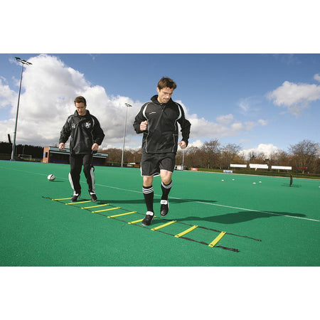 Two men in sportswear train on a green field, stepping through the Precision Training Adjustable Speed Ladder with adjustable rungs, placed on turf under a partly cloudy sky.