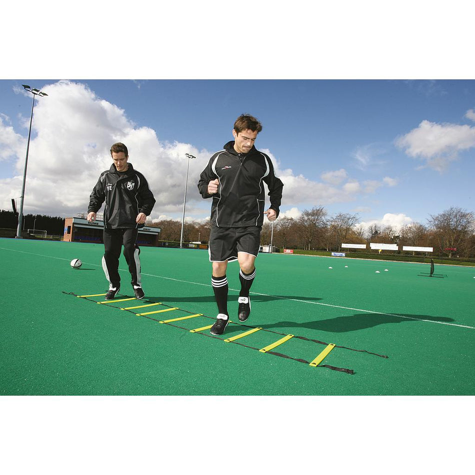 Two men in sportswear train on a green field, stepping through the Precision Training Adjustable Speed Ladder with adjustable rungs, placed on turf under a partly cloudy sky.