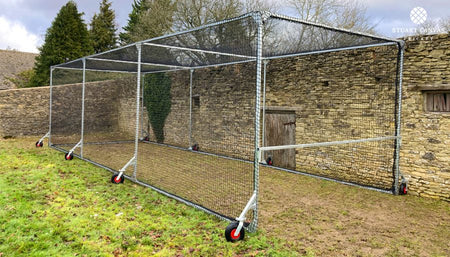 The Stuart Canvas Test Batting Cage is a mobile cricket enclosure with a metal frame, black mesh, and red-and-black wheels—perfect as a heavy-duty practice cage on grass near buildings or trees.