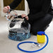 A person kneels on the floor, pouring water from a jug into a clear Urban Fitness Hydro Inertia Kettlebell, perfect for functional training. A blue and yellow air pump with hose is nearby on the carpet.