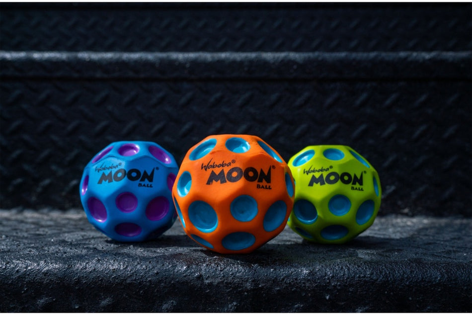 Three Waboba Martian Moon Balls—blue with purple, orange with blue, and green with blue—by Waboba sit side by side on textured black steps, each featuring large circular holes and designed for high bouncing fun.