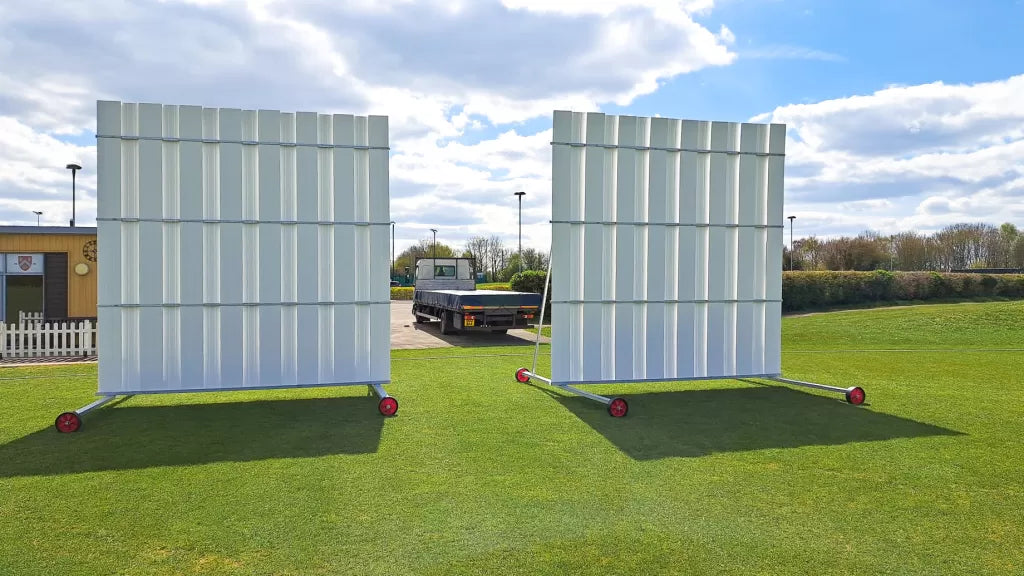 Two large Stuart Canvas Polypropylene Sightscreens on wheels stand on a green field, with a flatbed truck in the background beneath a partly cloudy sky.