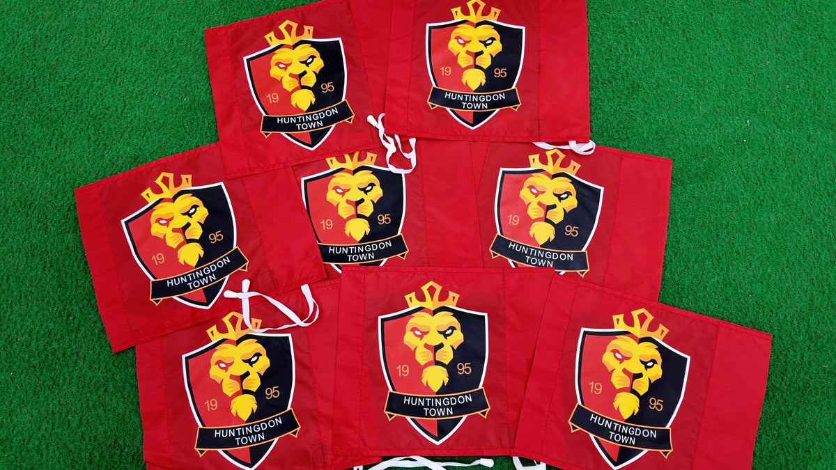 Seven custom printed corner post flags from Martin Berrill Sports, featuring a yellow lion emblem, crown, "HUNTINGDON TOWN" text, and the year 1995 in yellow and red, are displayed on green artificial grass. Perfect for personalisation.