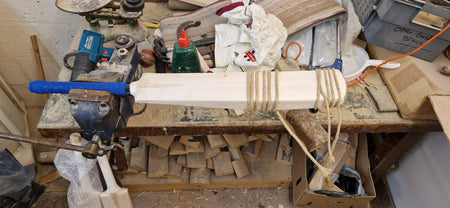 A Martin Berrill Sports Cricket Bat Repair - Toe, Shoulder, Edge is secured on a workbench with a blue grip, surrounded by tools and wood shavings—twine wrapped around the blade highlights the repair and restoration process in progress.