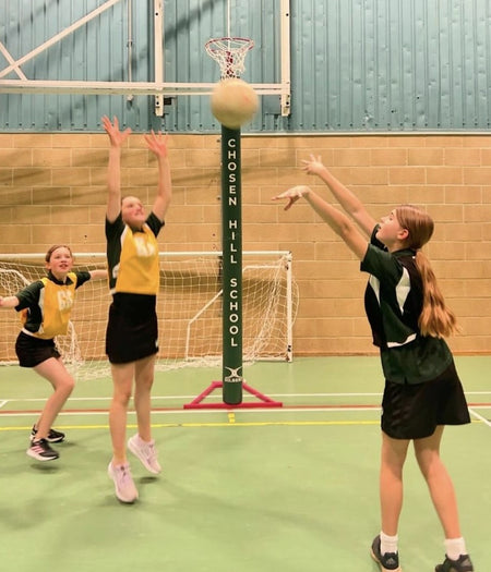 Three girls play netball at Chosen Hill School. One shoots while another blocks by Gilbert Custom Printed Netball Post Protectors (Pair)—personalised for your team—while a third player watches nearby.