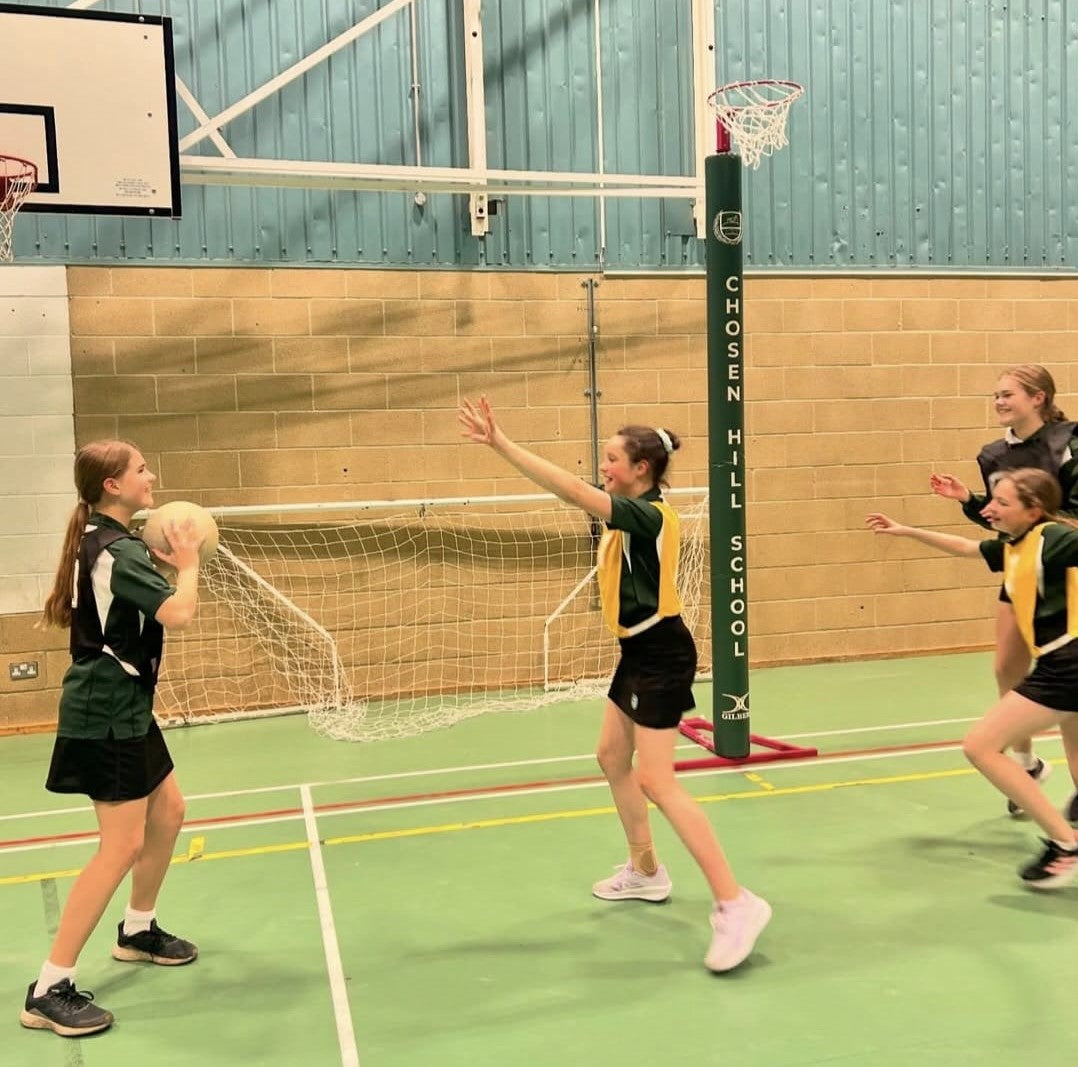 Four girls in sports uniforms play netball in a gym, with one holding the ball. A green Gilbert Custom Printed Netball Post Protector shows the Chosen Hill School name under the net.