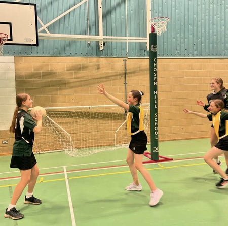Four girls in sports uniforms play netball in a gym, with one holding the ball. A green Gilbert Custom Printed Netball Post Protector shows the Chosen Hill School name under the net.