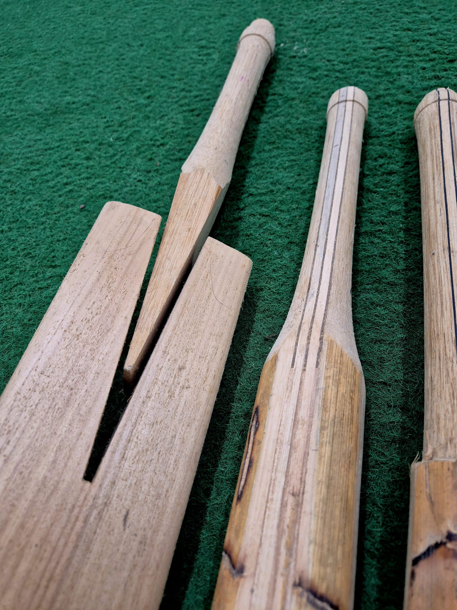Three wooden cricket bats lie on green turf: one with a large split down the blade—perfect for Cricket Bat Rehandle by Martin Berrill Sports—while the other two are intact but show signs of wear.