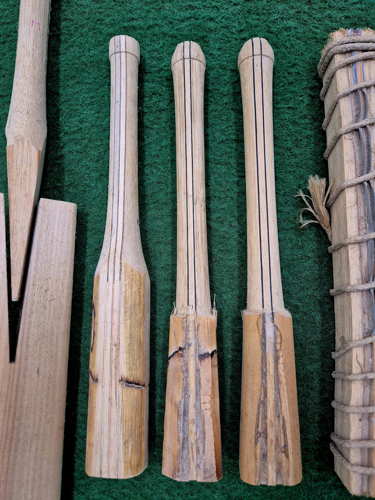 Three broken wooden handles with black lines, likely for a Cricket Bat Rehandle by Martin Berrill Sports, stand vertically on green textured fabric. Nearby are other wooden pieces and a twine-wrapped bundle, suggesting bat repair in progress.