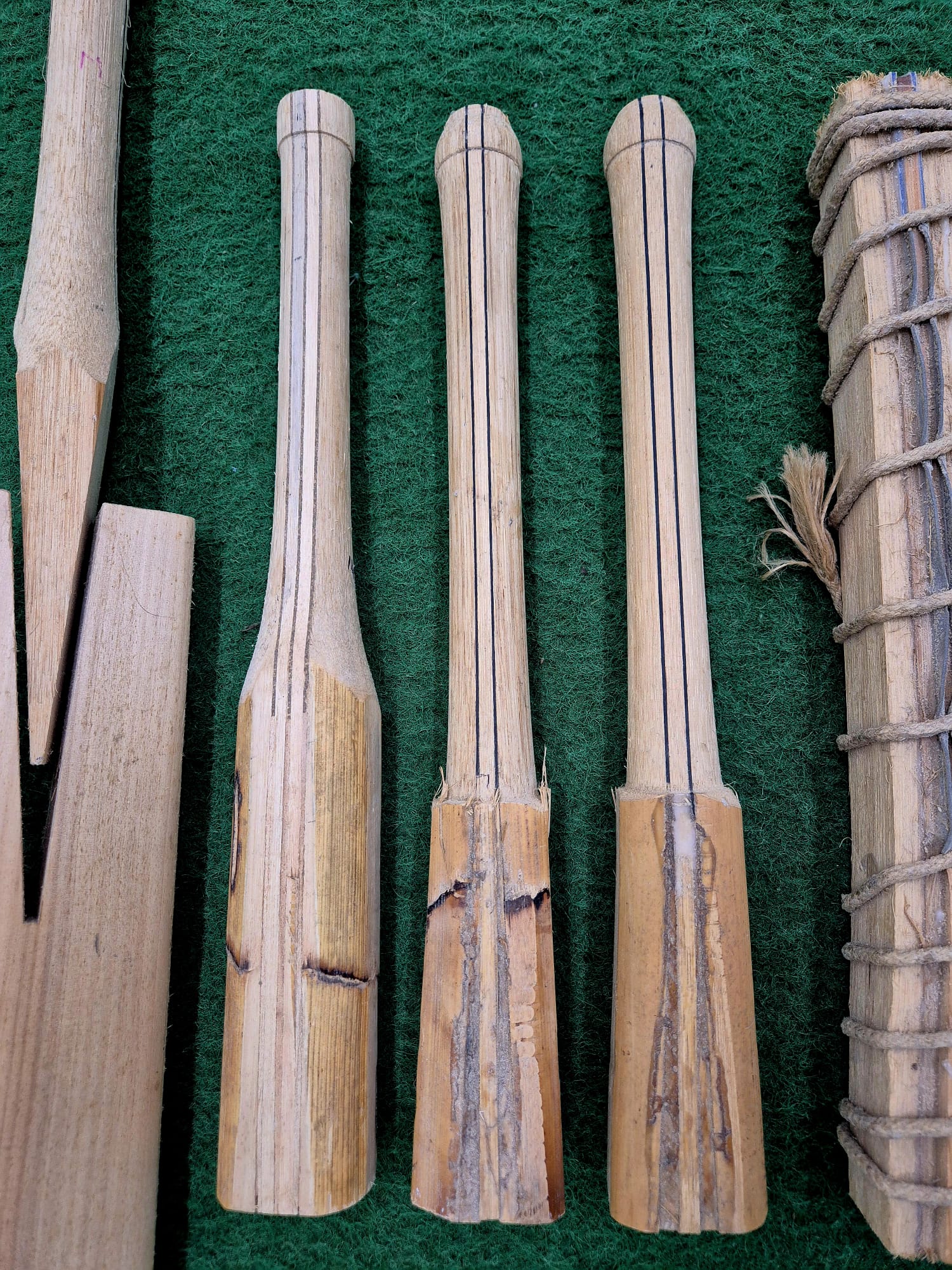Three broken wooden handles with black lines, likely for a Cricket Bat Rehandle by Martin Berrill Sports, stand vertically on green textured fabric. Nearby are other wooden pieces and a twine-wrapped bundle, suggesting bat repair in progress.