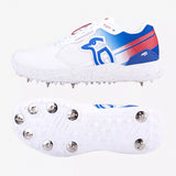 A pair of white Kookaburra KC 1.0 Cricket Spikes by Kookaburra, featuring blue and red accents, metal spikes on the sole, and a penguin logo on the side. The image shows a side view and the spiked sole.