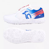 The Kookaburra KC 1.0 Rubber Soled Cricket Shoe by Kookaburra is a white high-performance cricket shoe with blue and red accents, branded side logo, and a textured sole with small spikes for grip on synthetic surfaces. Side and sole views shown.