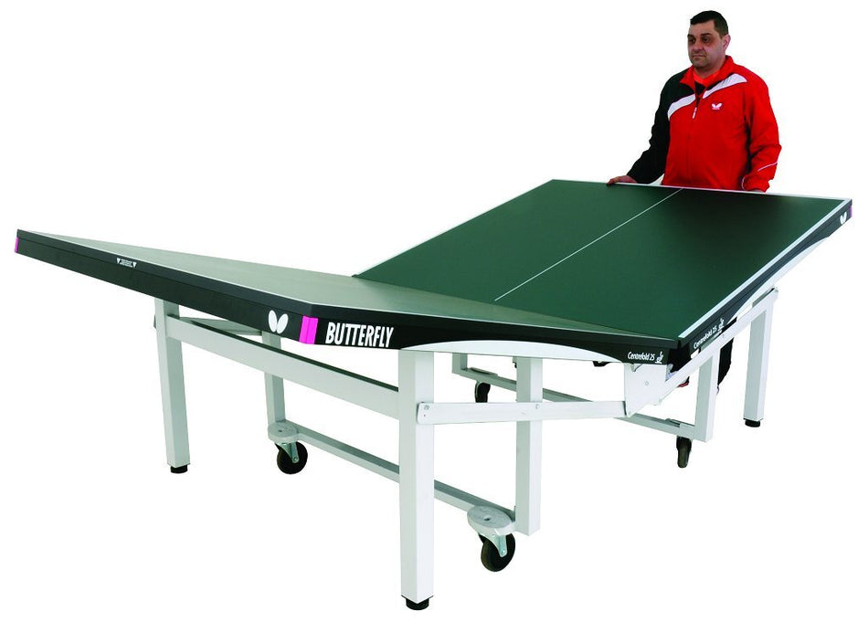 A man stands beside a tilted green Butterfly Centrefold 25 Rollaway Table Tennis Table, with one side raised and wheeled legs for easy movement; this ITTF approved model by Butterfly features a 25mm Anti Skid playing surface designed for optimal performance.