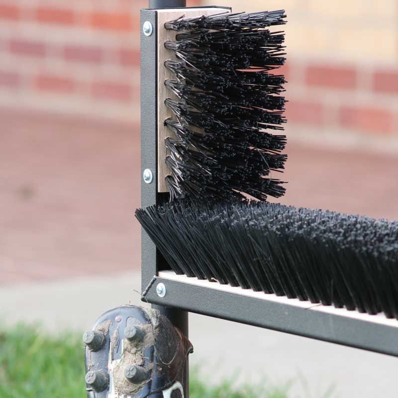 The Harrod Sport Multi Boot Wiper features stiff black bristles on a steel frame, ideal for cleaning boots or shoes. Shown close-up with a blurred brick wall and green grass in the background.