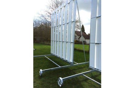 The Martin Berrill Sports Club PVC Sight Screen, a large metal A-frame structure with wheels for mobility, stands on a grassy lawn. Trees and a house are visible in the background beneath a cloudy sky.