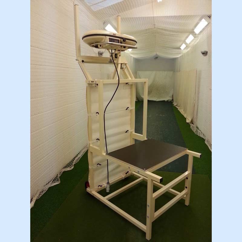The Bola Bowling Machine Stand on Wheels by Bola, equipped with a seat and desk, is set up in an indoor net area featuring green artificial turf flooring and white netted walls.