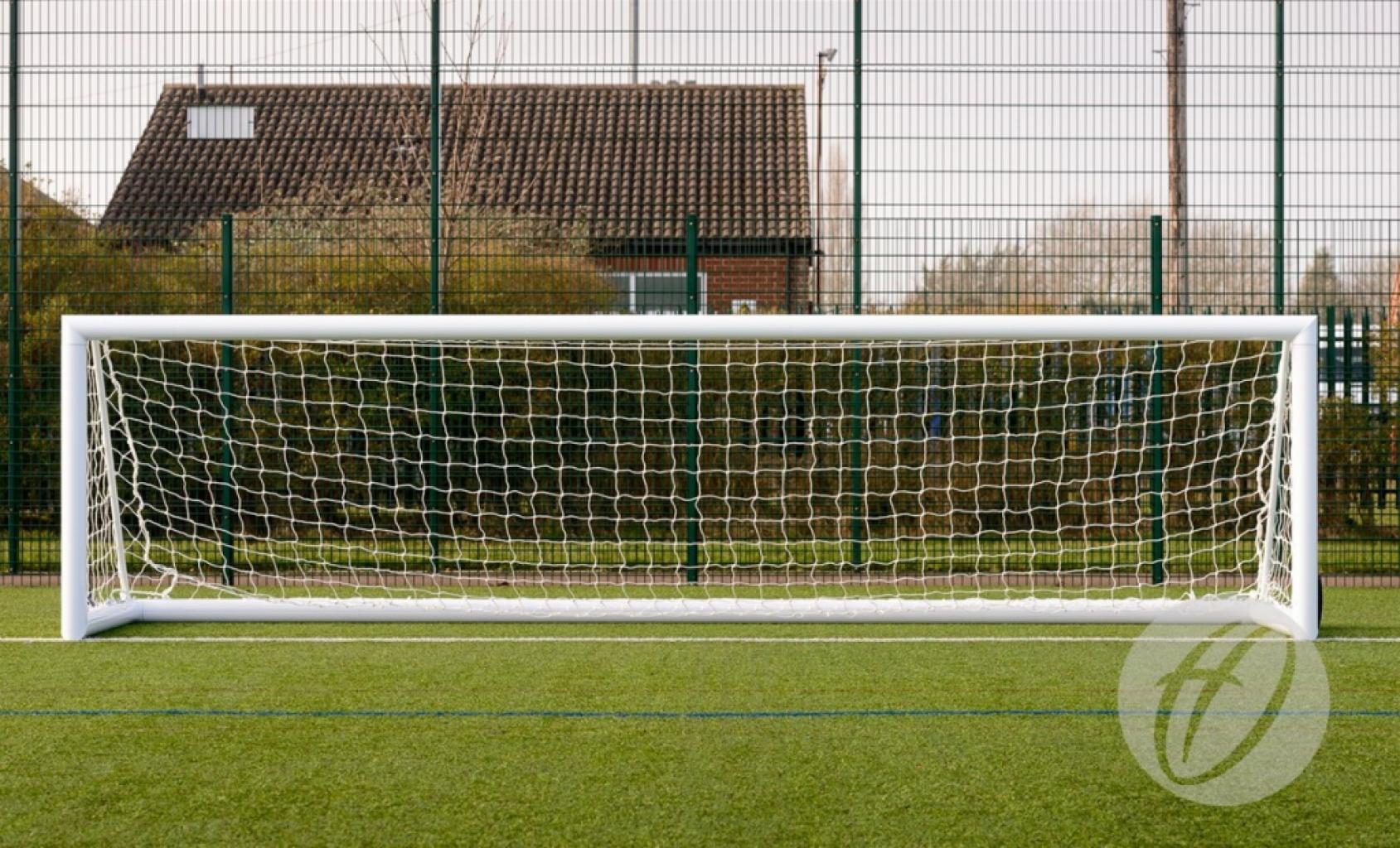 A Harrod Sport 4G Weighted Portagoal – 5v5, measuring 4.88 x 1.22M, stands on green artificial turf, with a metal fence, bushes, and a building with a tiled roof visible in the background.