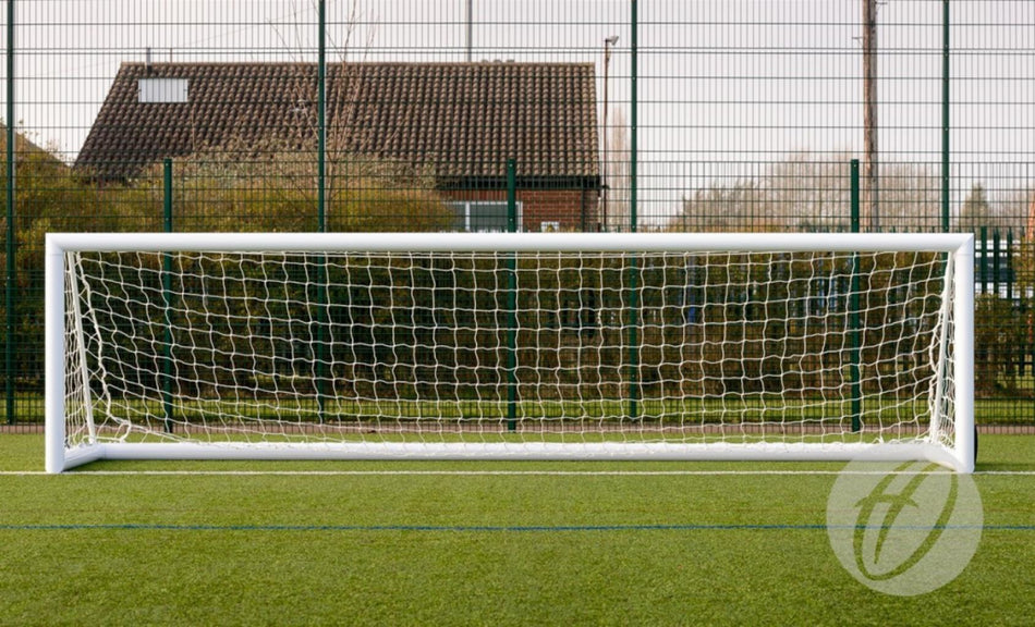 A Harrod Sport 4G Weighted Portagoal – 5v5, measuring 4.88 x 1.22M, stands on green artificial turf, with a metal fence, bushes, and a building with a tiled roof visible in the background.