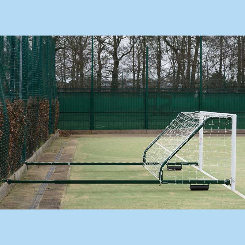 The Harrod Sport 4G Fence Folding Goal (3.66m x 1.22m, 2.3m to 3.5m proj) is tipped forward on a fenced outdoor field, showcasing its reinforced aluminium side bars and durable netting. Backed by tall green fencing and trees, this goal is ideal for synthetic surfaces. The image features a light blue border.