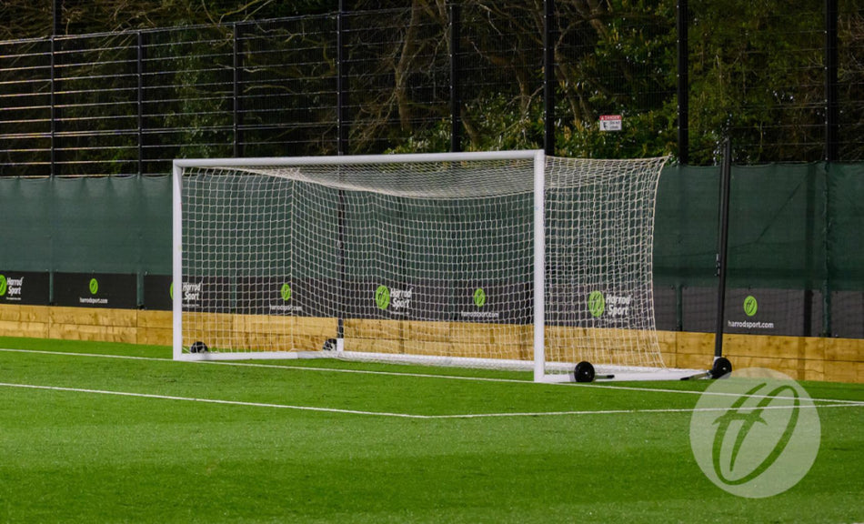 A Harrod Sport Weighted UEFA Portagoal stands on a green artificial turf field, with netting behind it and a fence in the background—resembling the classic stadium football goal design.